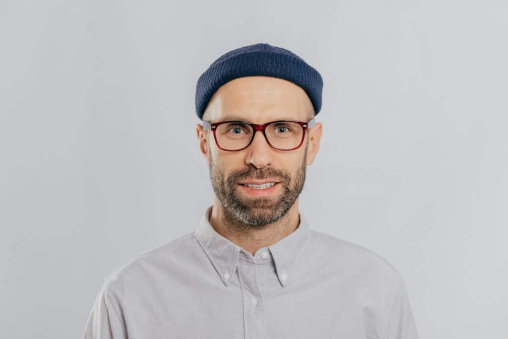 Небритый мужчина в прозрачных очках, головном уборе, формальной рубашке, смотрящего прямо в камеру, изолированный на белом фоне