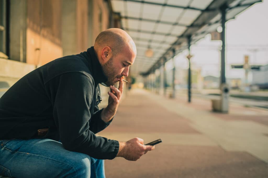 Uomo pelato fuma una sigaretta su una panchina mentre usa il suo smartphone