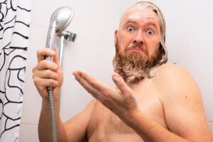 Homme barbu, perplexe, savonneux et mouillé, debout dans une baignoire et tenant une pomme de douche à la main.